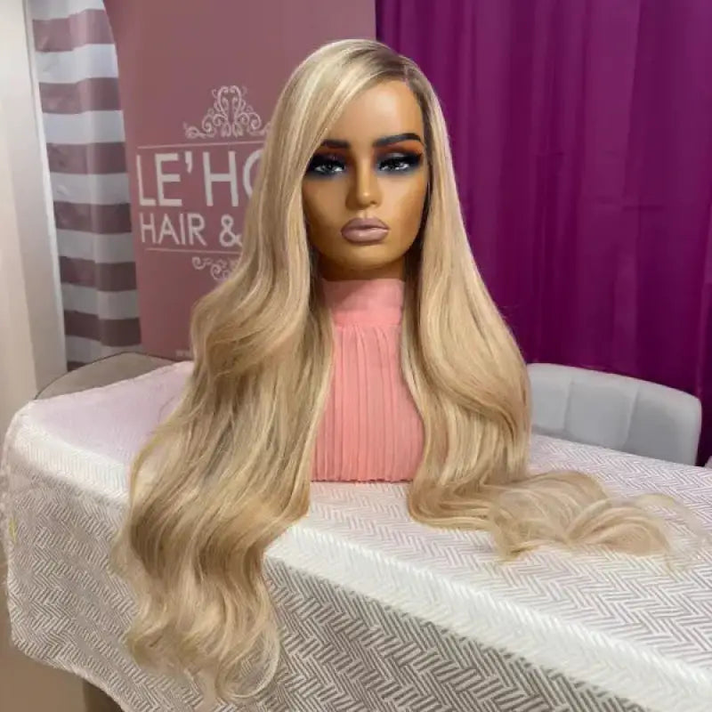 Blonde wig on a mannequin head with a pink top, placed on a table with a purple curtain in the background.